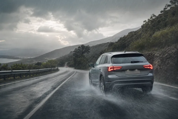 Turismo circulando despacio sobre una carretera mojada bajo lluvia, con las luces encendidas y gran separación respecto al coche precedente